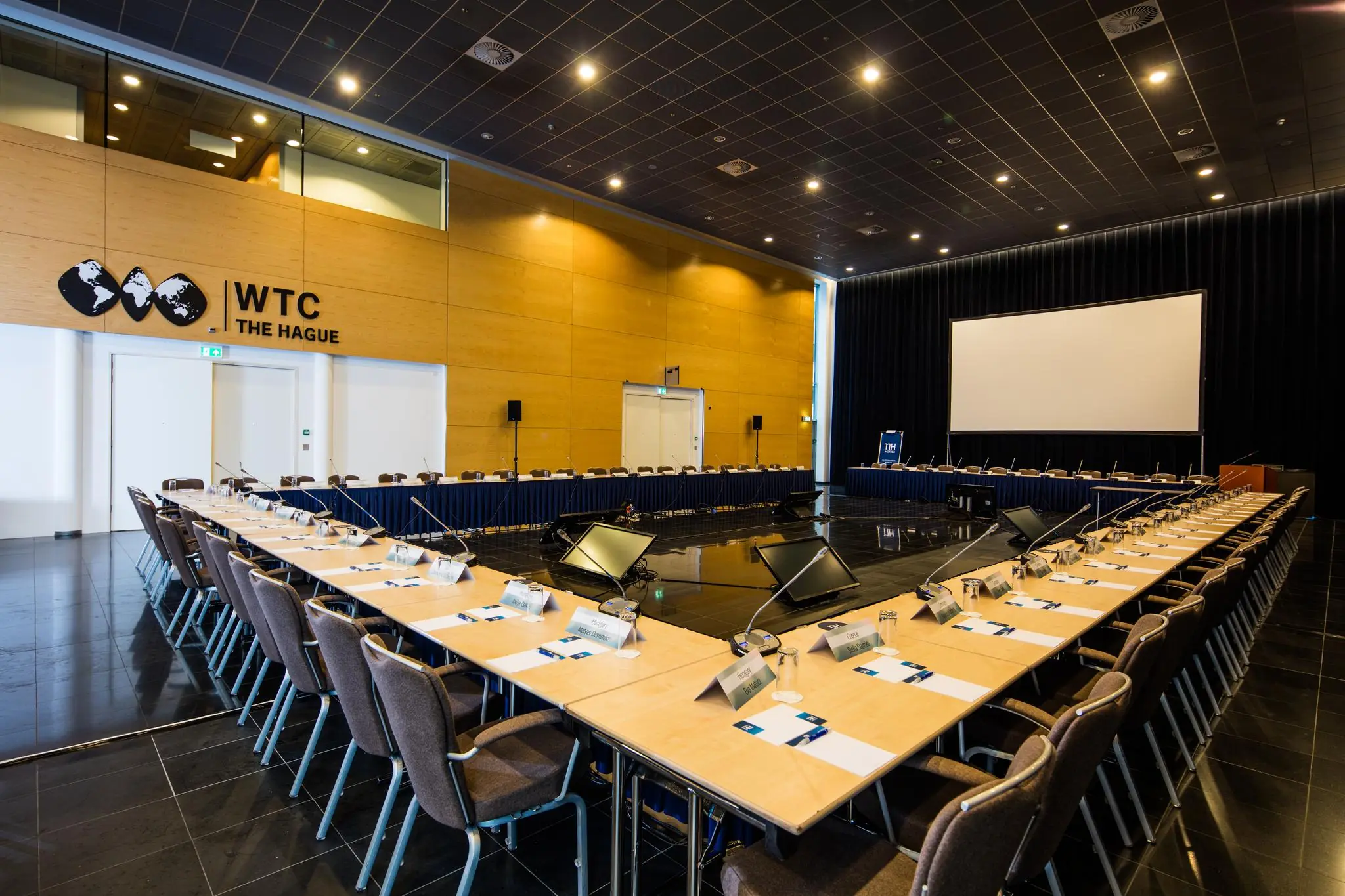 Vergaderzaal in het WTC The Hague aan de Prinses Beatrixlaan, ingericht voor een conferentie met microfoons, naamkaartjes en groot projectiescherm.